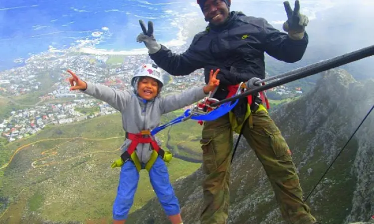 Man and child secured by ropes, smiling and throwing peace signs with a townscape in the background.