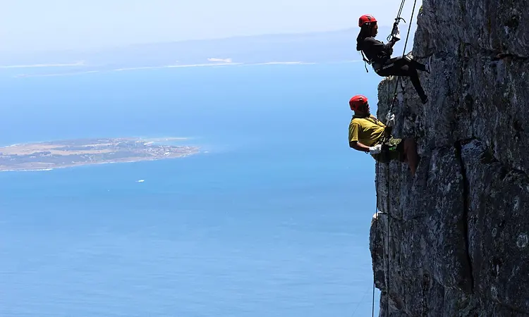 Two people abseiling down a cliff face with the ocean stretching out behind them.