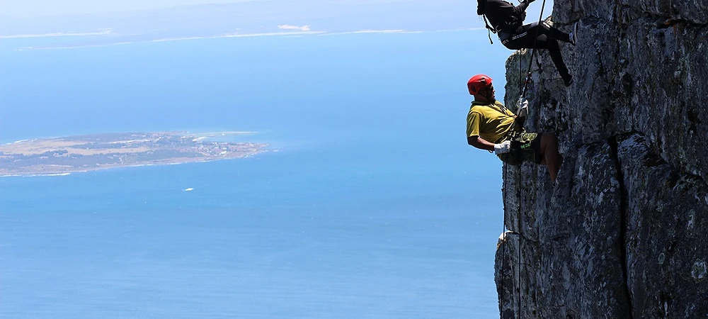 Two people abseiling down a cliff face with the ocean stretching out behind them.